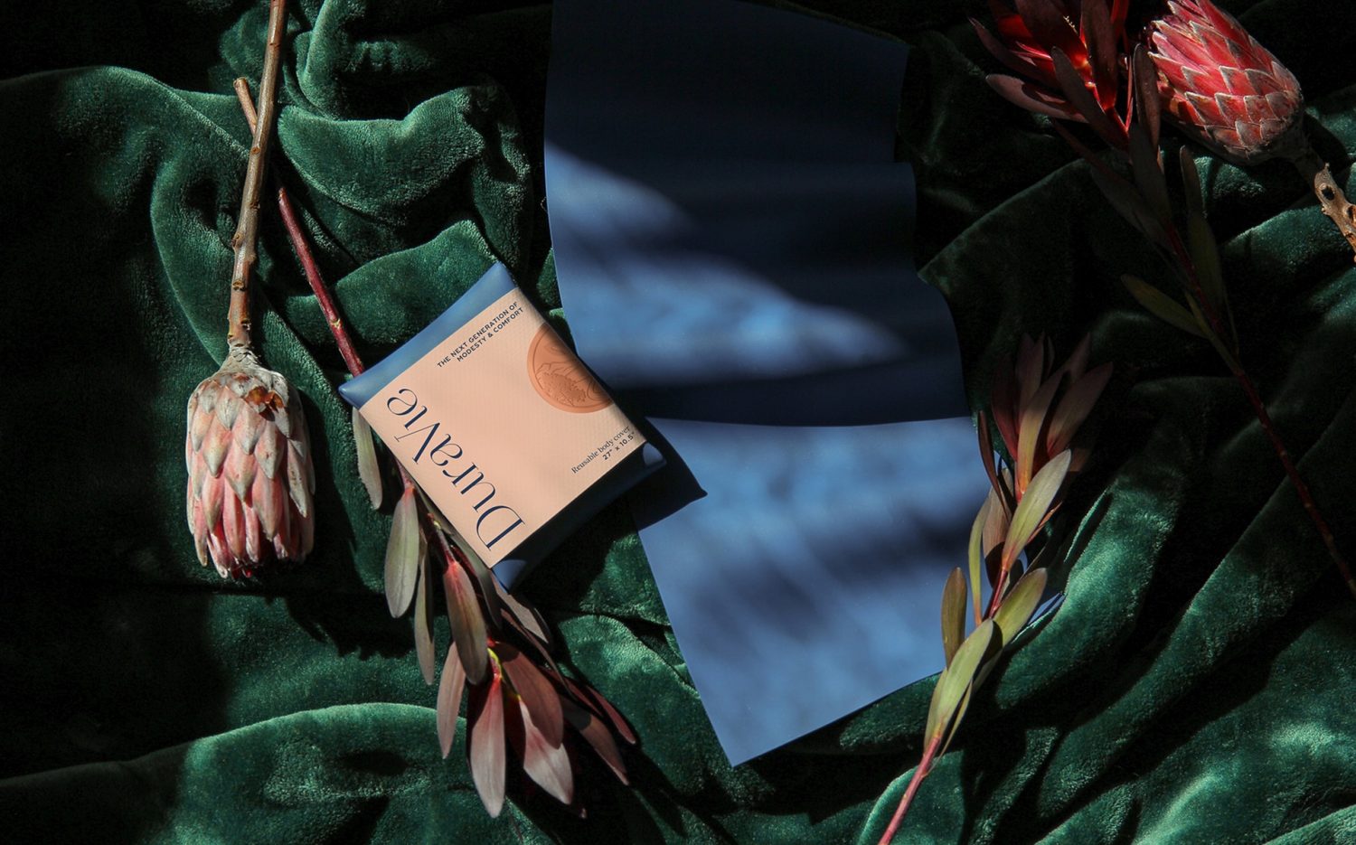 Duravie body wrap folded with newly branded identity label. It lays on green velvet with an unfolded body wrap. Protea flowers surround the products.