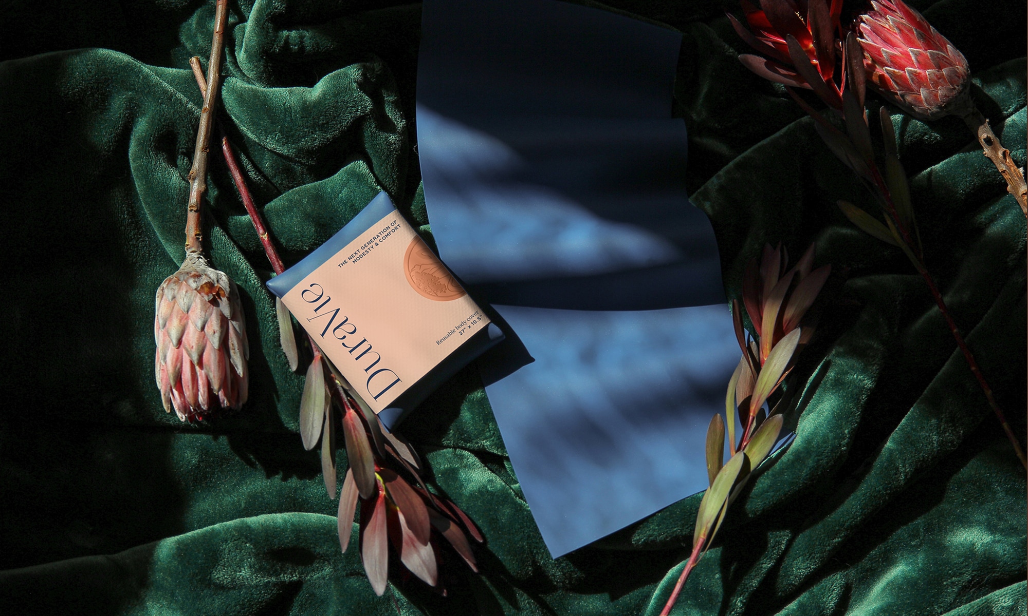Duravie body wrap folded with newly branded identity label. It lays on green velvet with an unfolded body wrap. Protea flowers surround the products.