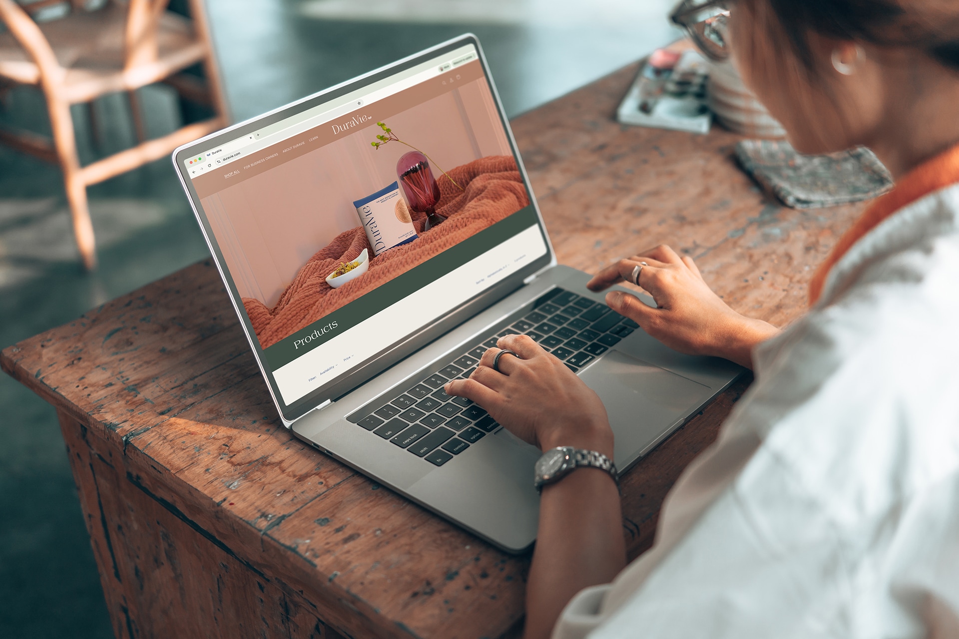 A woman on her laptop viewing the DuraVie product page. The colors are warm terracottas with a wooden desk.